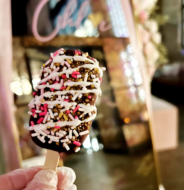 Hand holding a chocolate-dipped ice cream bar with white drizzle and crunchy topping
